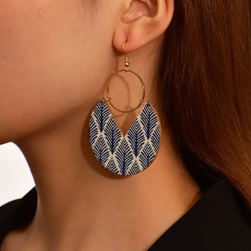 Close-up of a woman wearing a single bohemian wooden drop earring with a blue leaf pattern and gold-tone hoop.