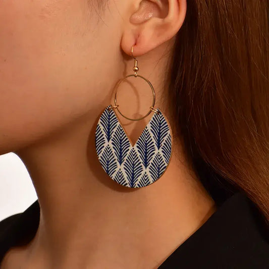 Close-up of a woman wearing a single bohemian wooden drop earring with a blue leaf pattern and gold-tone hoop.