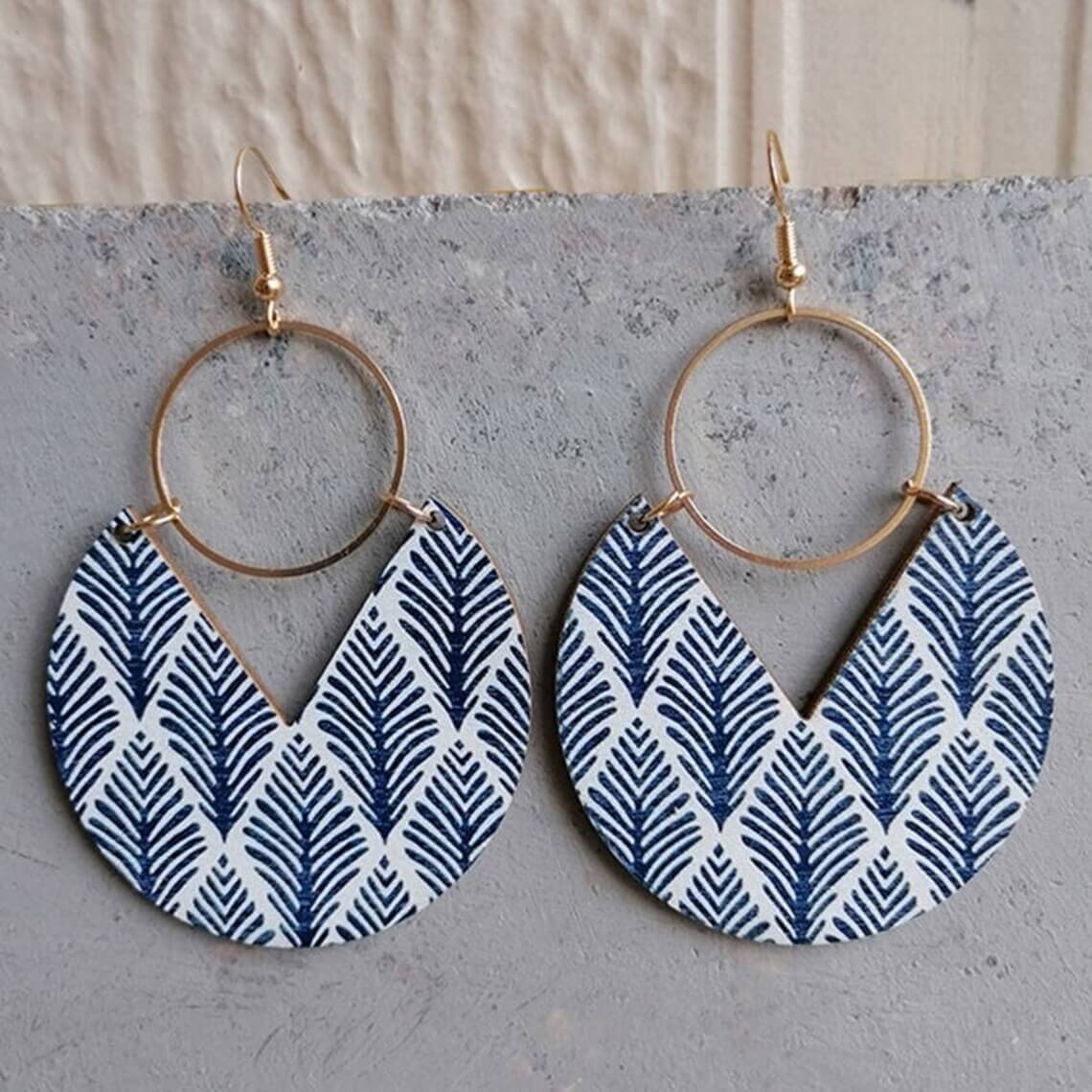 Pair of bohemian wooden drop earrings with blue leaf pattern and gold-tone hoops, displayed on a concrete surface.