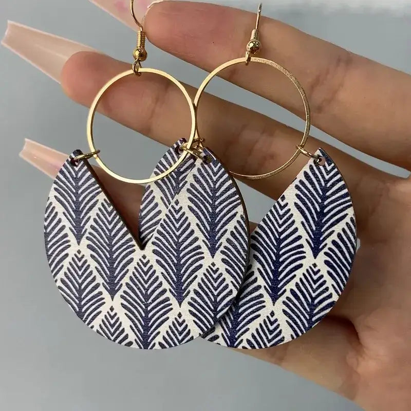 Hand holding a pair of bohemian wooden drop earrings with blue leaf pattern and gold-tone hoops.