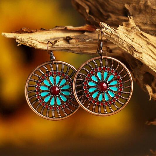 A pair of boho earrings with a mandala design, turquoise petals, and red accents, hanging from a tree branch against a blurred warm background.