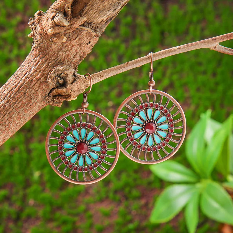 A pair of boho earrings with a mandala design, turquoise petals, and red accents, hanging from a tree branch against a vibrant green, mossy background.