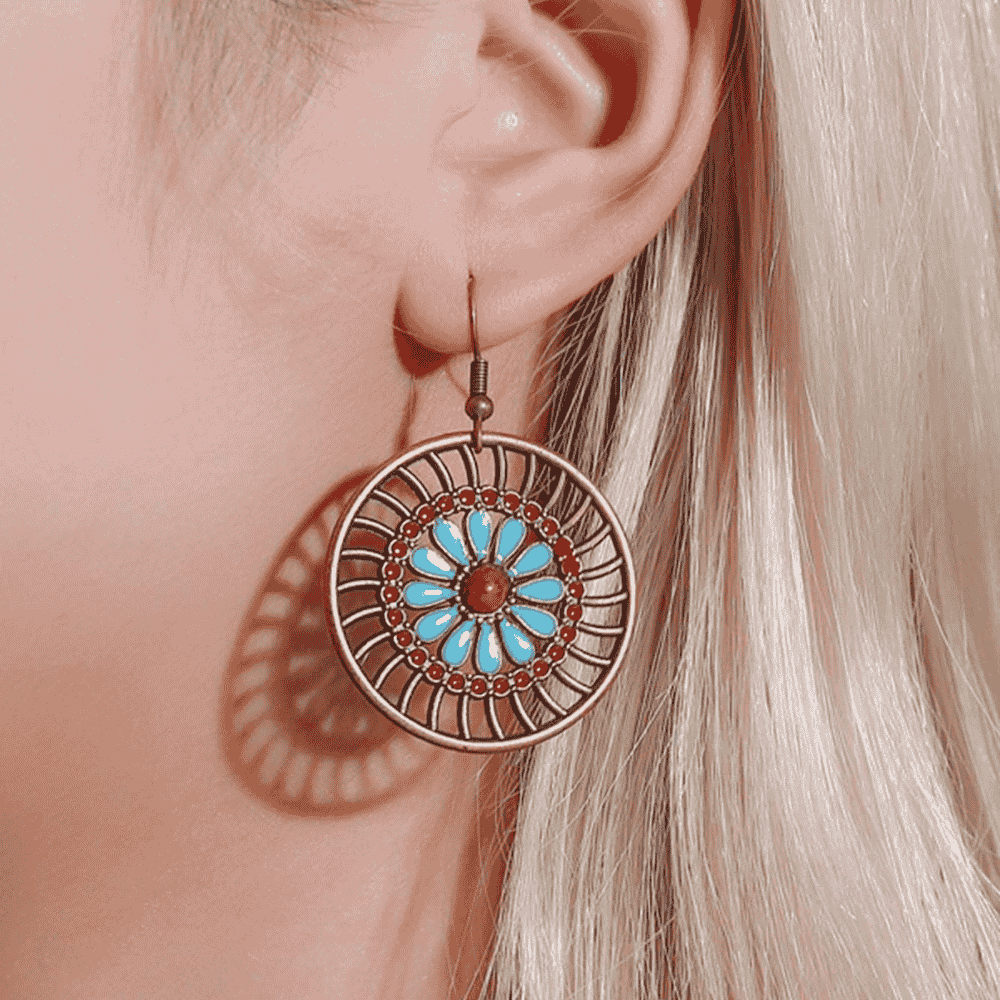 A close-up of a woman's ear wearing a single boho earring with a mandala design, turquoise petals, and red accents, against her blonde hair.