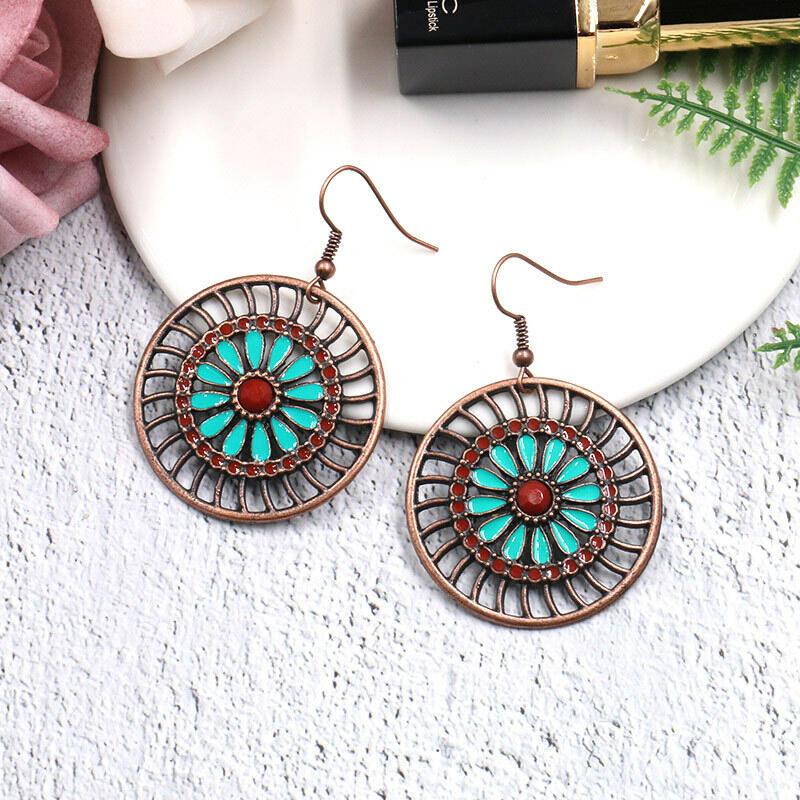 A single boho earring with a mandala design, turquoise petals, and red accents, hanging from a tree branch against a blurred natural background.