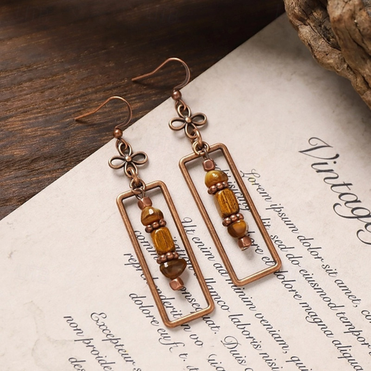 A pair of boho earrings with copper rectangular frames and brown and gold beaded accents, resting on a vintage-style paper.