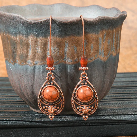 A pair of boho copper drop earrings featuring a teardrop shape with a brown stone, intricate filigree details, and small wooden beads on the ear wire, set against a rustic ceramic pot.