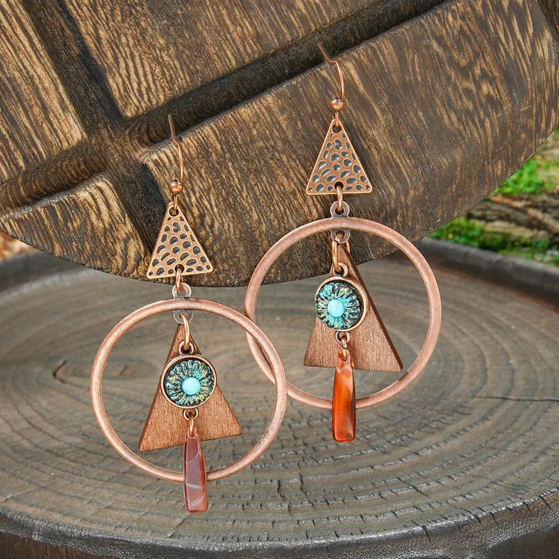 A pair of boho earrings with copper geometric shapes, wooden triangles, turquoise centers, and orange stone drops, displayed on a wooden surface.