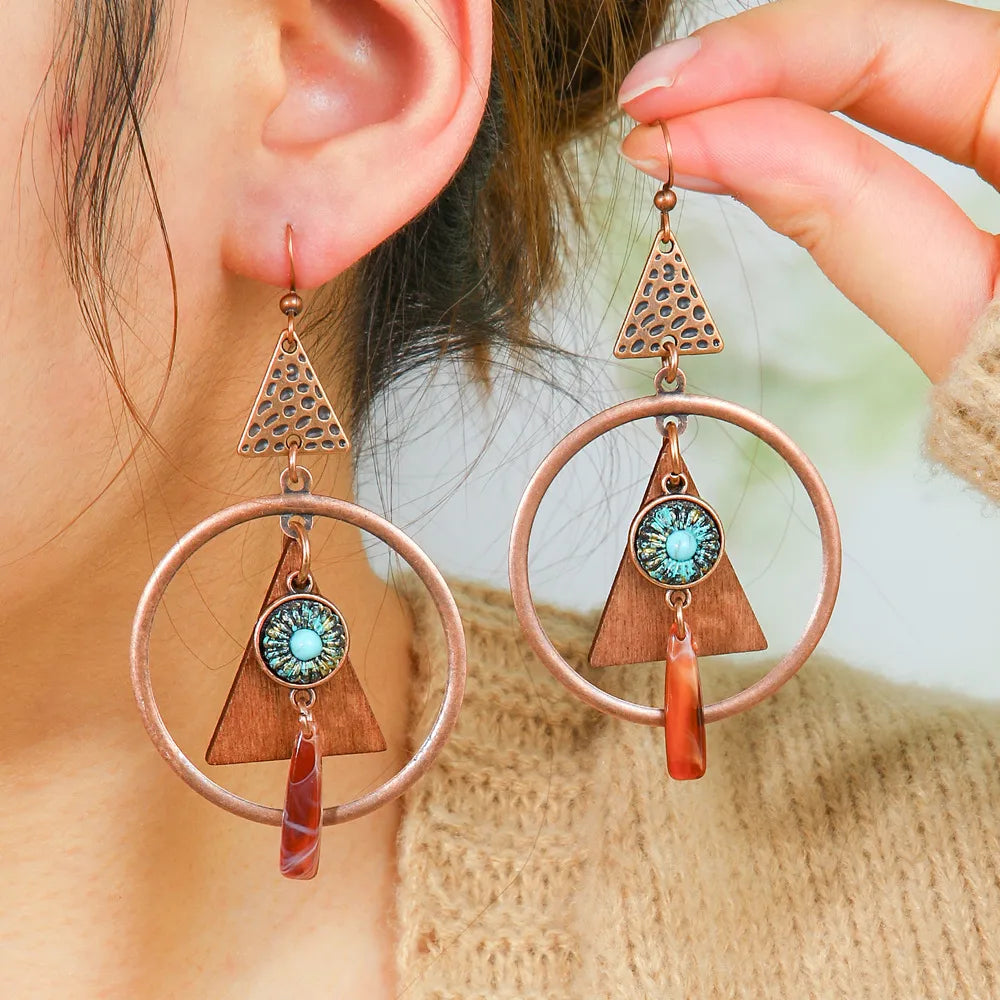 A model wearing a pair of long boho earrings featuring copper geometric elements, wooden triangles, turquoise details, and orange stone drops.