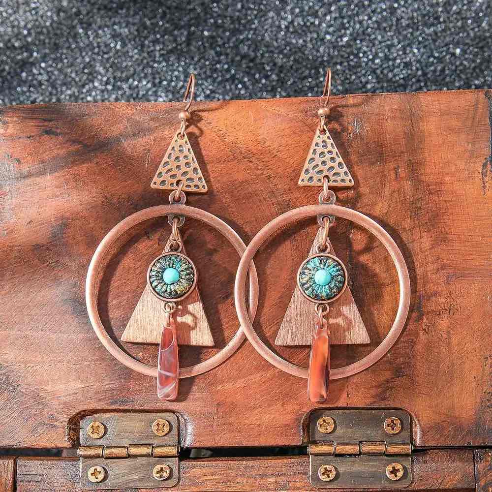 A pair of boho earrings with copper geometric shapes, wooden triangles, turquoise centers, and orange stone drops, laid on a rustic wooden box.