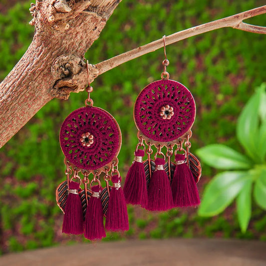 A pair of maroon crochet round tassel earrings with copper accents, hanging from a tree branch against a green background, showcasing their bohemian style.