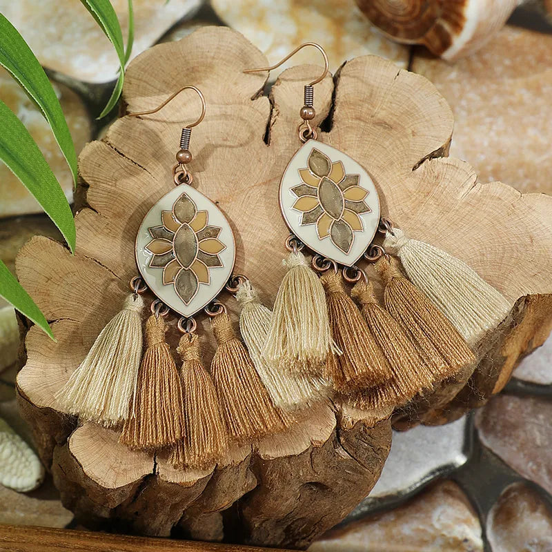 A pair of boho earrings with cream enamel, brown floral design, and layered beige and brown tassels, displayed on a wooden slab.