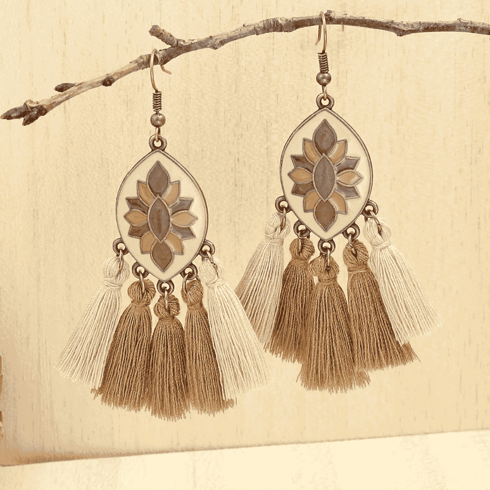 A pair of boho earrings with cream enamel, brown floral design, and layered beige and brown tassels, hanging from a bare tree branch against a light background.