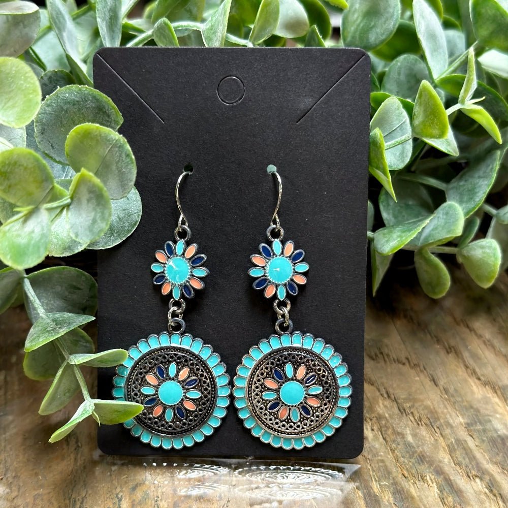 A pair of boho earrings with floral designs in turquoise, coral, and navy enamel, hanging from silver hooks on a black display card, with green foliage in the background.
