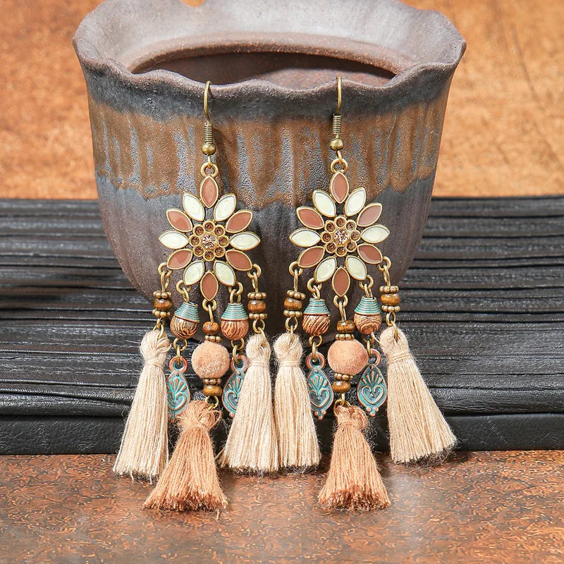A pair of long boho earrings with floral tops, beads, and multiple tassels in earthy brown and beige, displayed against a rustic background.