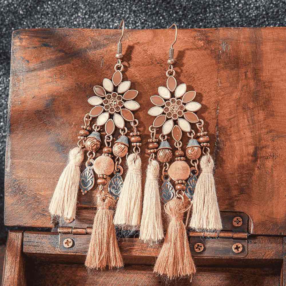 A pair of long boho earrings with floral tops, beads, and multiple tassels in earthy brown and beige, laid on a wooden box.