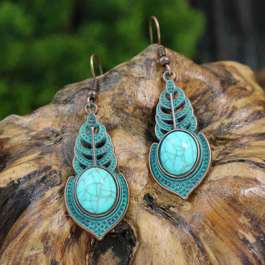 A pair of boho earrings with turquoise stones and an antique copper leaf design, displayed on a textured wooden surface.