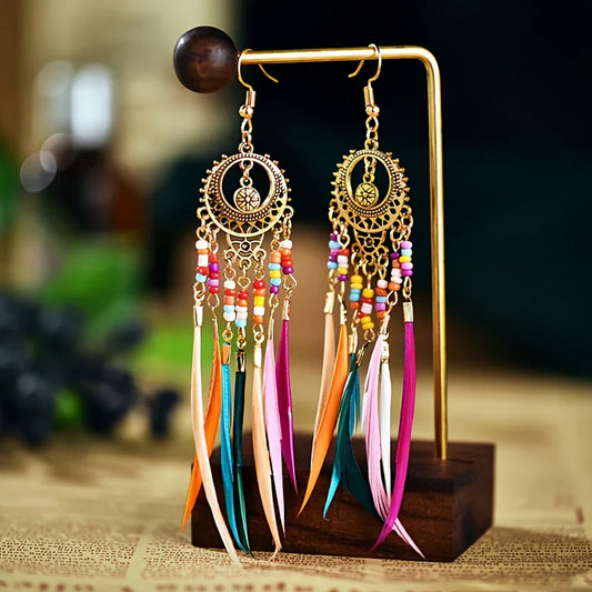 A pair of gold-toned metal boho earrings with colorful beads and long feather tassels, displayed on a wooden stand.