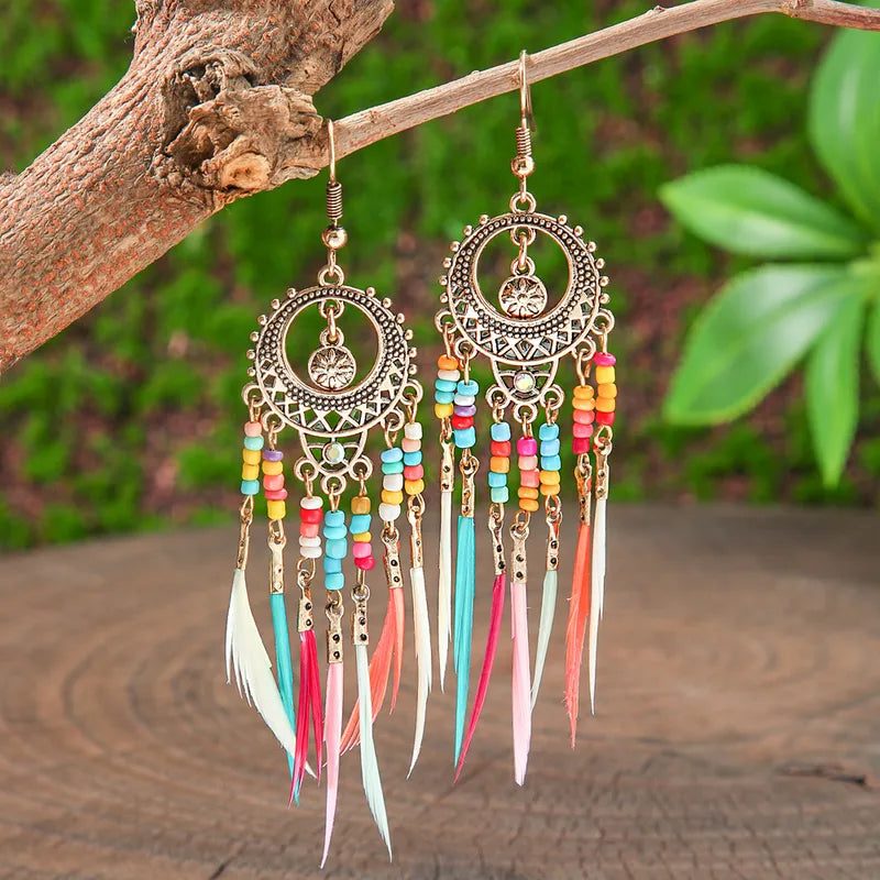 A pair of gold-toned metal boho earrings with colorful beads and long feather tassels, hanging from a tree branch.