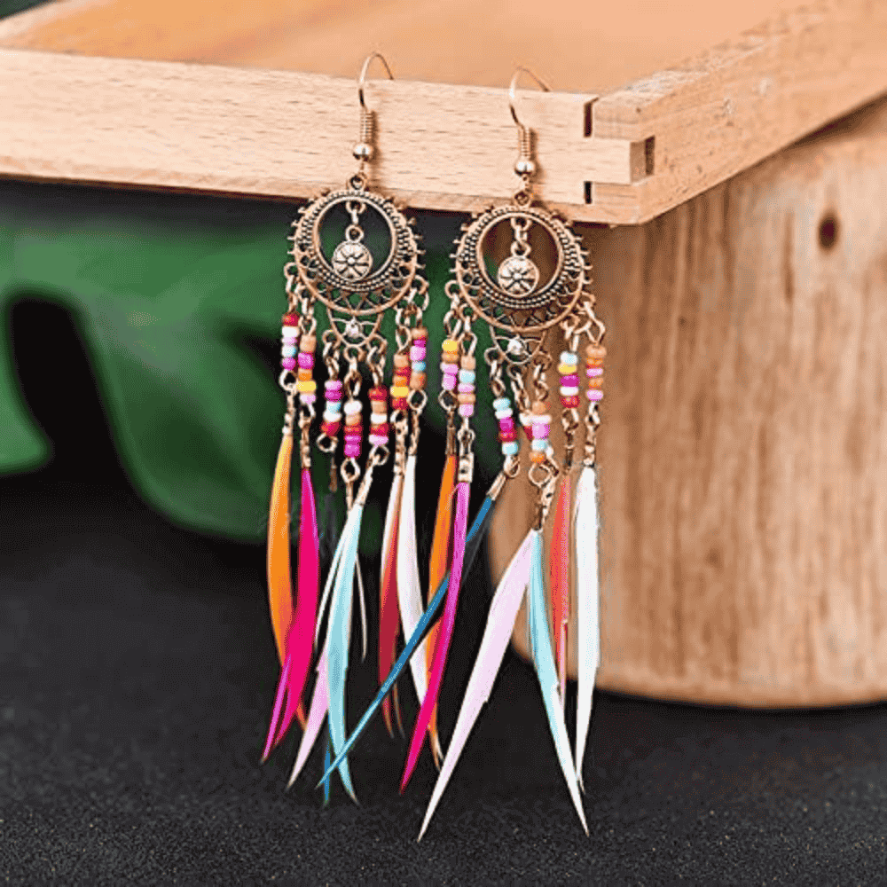 A pair of gold-toned metal boho earrings with colorful beads and long feather tassels, placed on a wooden box.