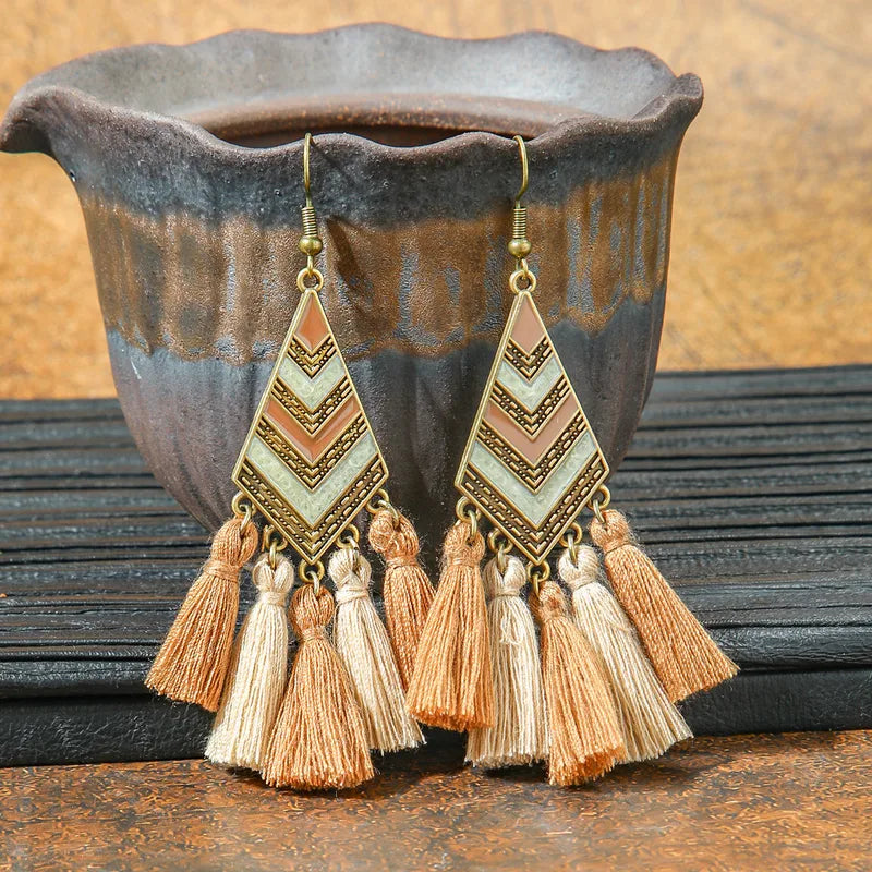 A pair of boho earrings with a bronze-toned metal geometric top featuring a beige and brown chevron pattern, and multiple fabric tassels in varying shades of brown and beige, displayed against a rustic ceramic pot.