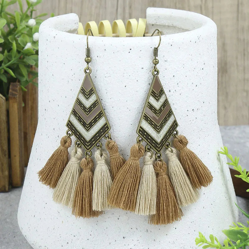 A pair of boho earrings with a bronze-toned metal geometric top featuring a beige and brown chevron pattern, and multiple fabric tassels in varying shades of brown and beige, displayed against a white textured background with green foliage.