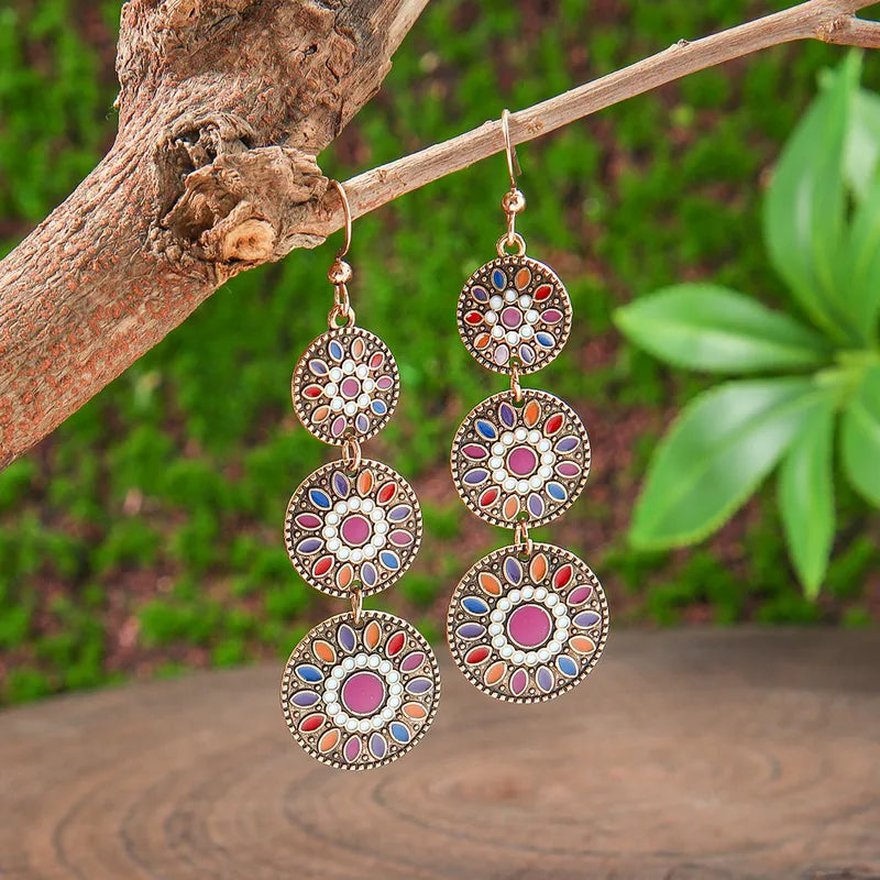 Close-up of two long boho earrings with multicolored mandala patterns, hanging from a tree branch against a blurred green background.