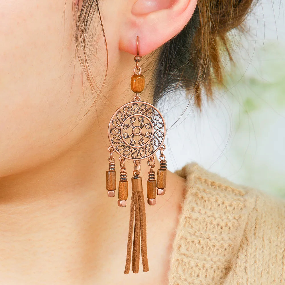 A woman wearing a single boho earring with an oxidised circular filigree design, wooden beads, and faux suede tassels, against a blurred background.