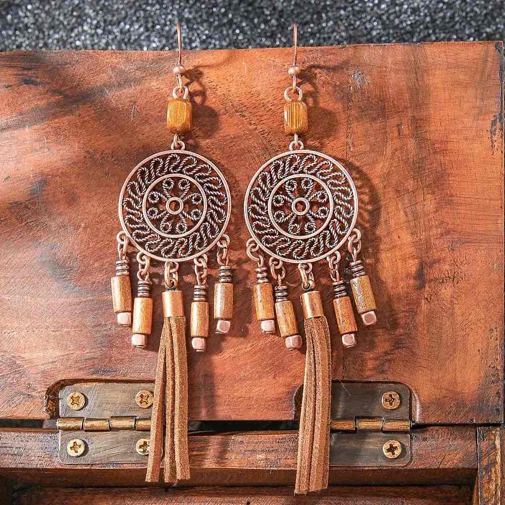 A pair of boho earrings with oxidised circular filigree design, wooden beads, and faux suede tassels, displayed on a wooden chest.