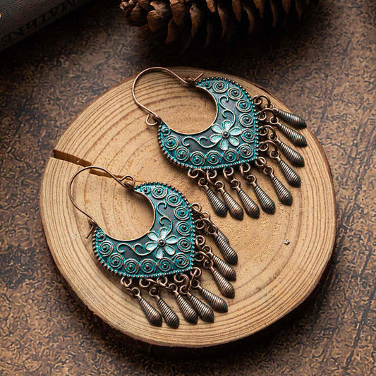 A pair of boho oxidised earrings with teal floral and swirl patterns and bronze dangling elements, displayed on a wooden surface.