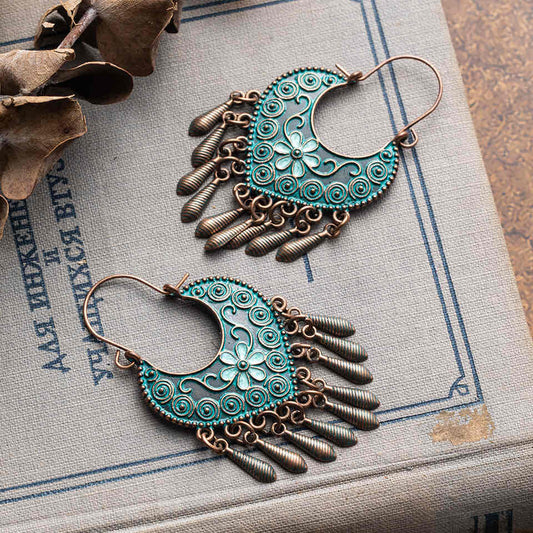 Two boho oxidised earrings with teal floral and swirl designs and bronze dangle accents, resting on a vintage book with Russian text.