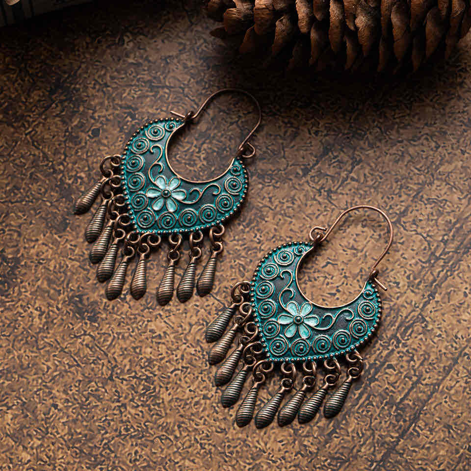 A pair of boho oxidised earrings featuring teal floral and swirl motifs and bronze tassels, set against a textured brown surface with a pine cone.