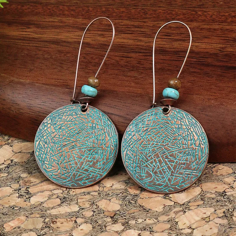 A pair of boho earrings featuring round oxidised alloy discs with a turquoise patina and small turquoise and brown beads, displayed on a wooden surface.