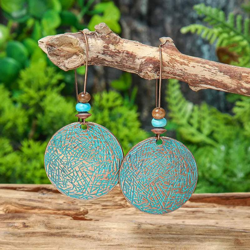 A pair of boho earrings with round oxidised alloy discs and turquoise and brown beads, hanging from a tree branch against a blurred green background.