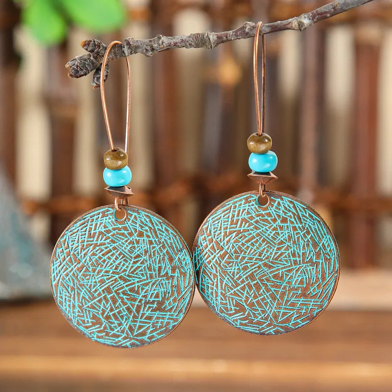 A pair of boho earrings featuring round oxidised alloy discs with a turquoise patina and small turquoise and brown beads, hanging from a tree branch.