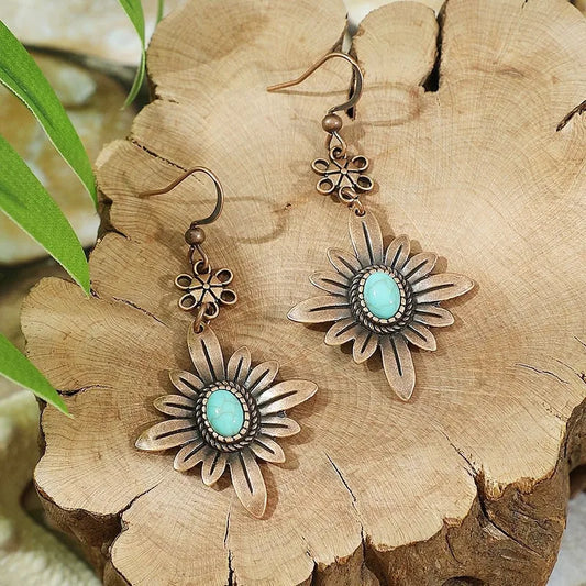 A pair of boho oxidised floral dangle earrings with turquoise stones, displayed on a wooden surface with green leaves in the background.