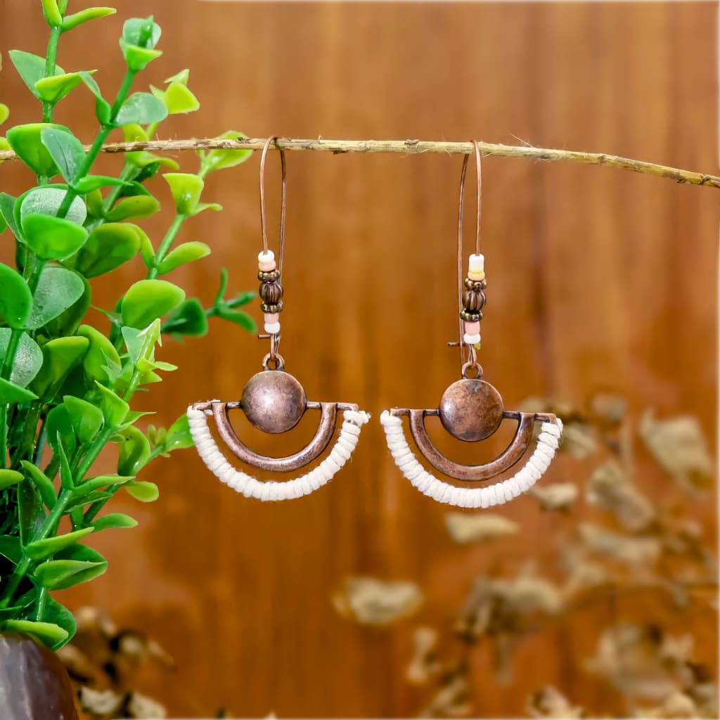 A pair of boho earrings, featuring oxidised half-moon shapes with white thread accents and small beads, hanging from a green leafy branch against a wooden background.