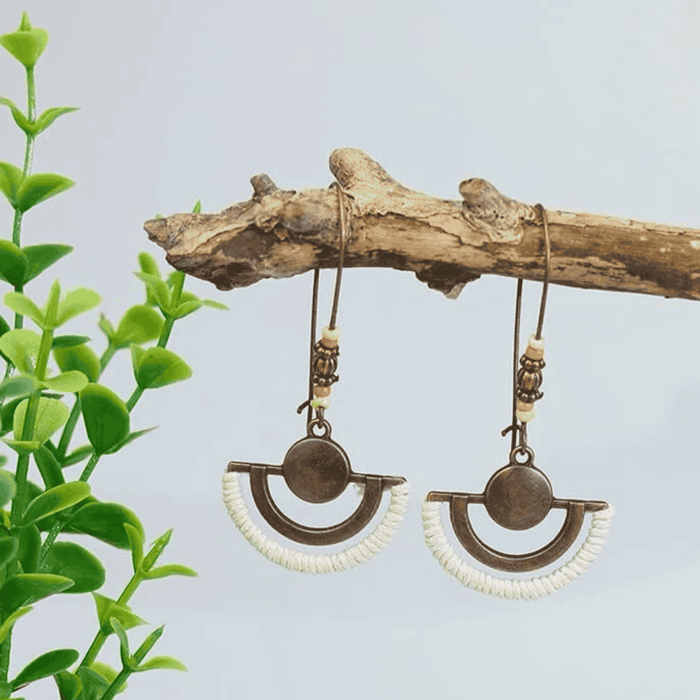 A pair of boho earrings with oxidised half-moon elements wrapped in white thread and small beads, suspended from a rustic wooden branch with green foliage in the background.