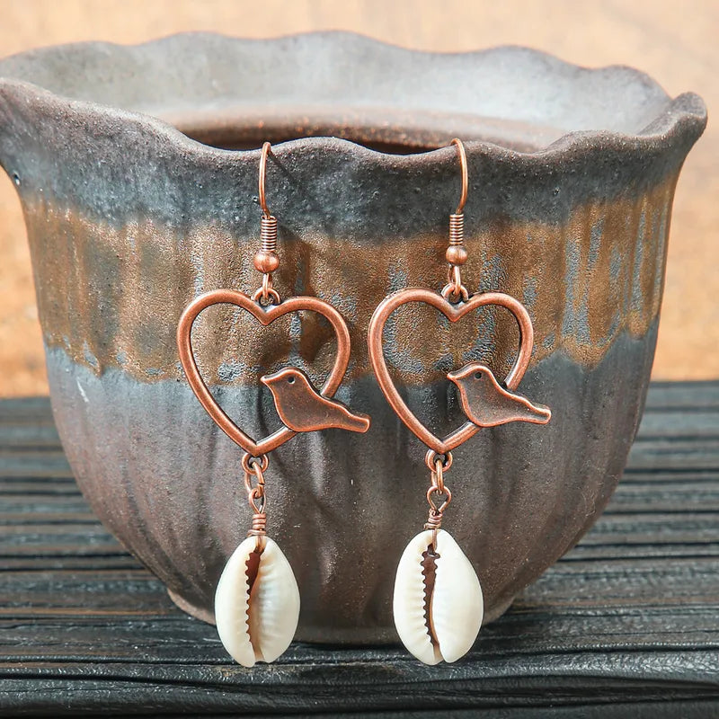 Close-up of a pair of boho earrings with oxidised bronze hearts, small birds, and white shell drops, resting in a rustic ceramic pot.