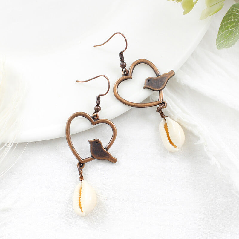 A pair of boho earrings with oxidised bronze heart and bird designs, and white shell drops, displayed on a white surface with a hint of green foliage.