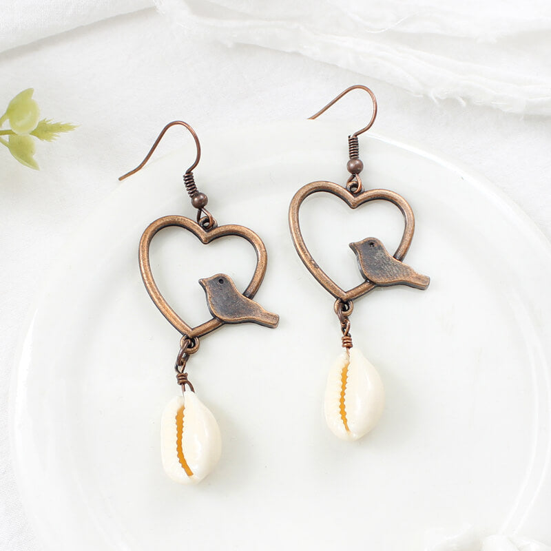 Two boho earrings featuring oxidised bronze hearts, small birds, and white shell drops, neatly arranged on a white plate.