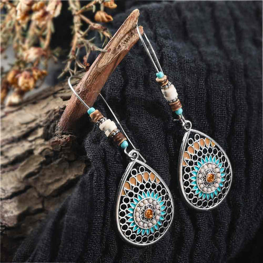A pair of boho earrings with oxidised silver teardrop mandalas and beaded hooks, displayed on a dark fabric next to a wooden branch.