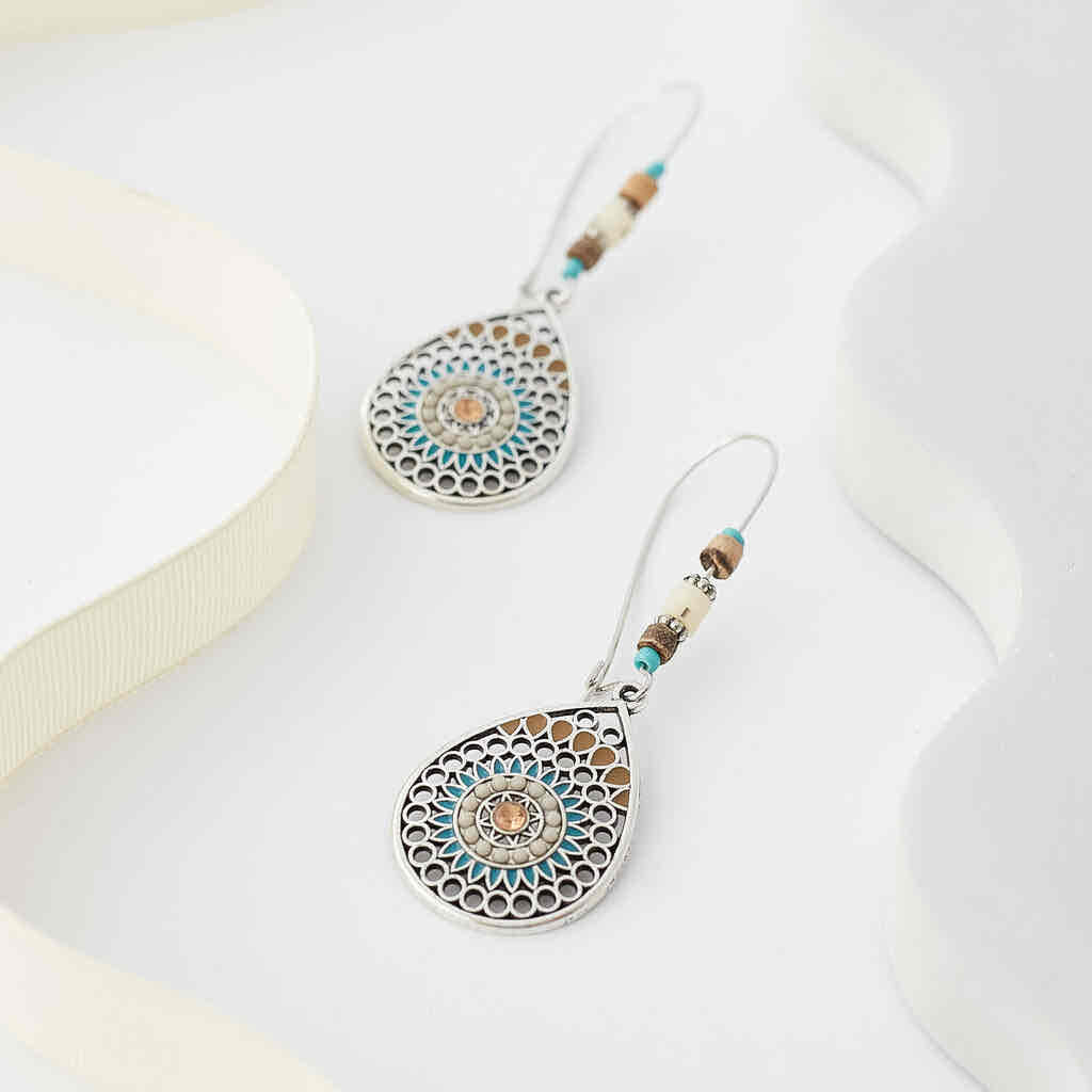A pair of boho earrings with oxidised silver teardrop mandalas and beaded hooks, displayed on a dark fabric next to a wooden branch.