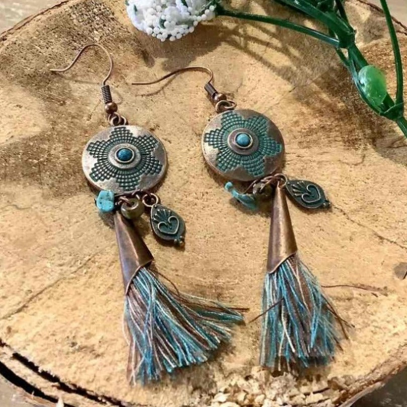 Close-up of a pair of oxidised metal boho earrings with turquoise accents and blue-green tassels, on a wooden background.