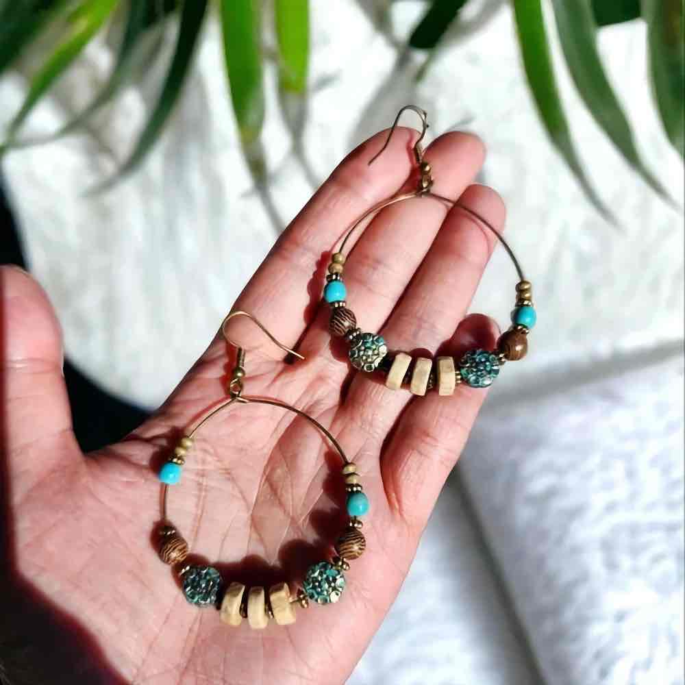 A hand holding a pair of large boho hoop earrings adorned with turquoise and wooden beads, against a soft background.