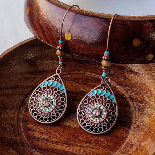 Close-up of a pair of boho earrings with oxidised metal teardrop pendants featuring a mandala pattern and turquoise accents, resting in a wooden bowl.