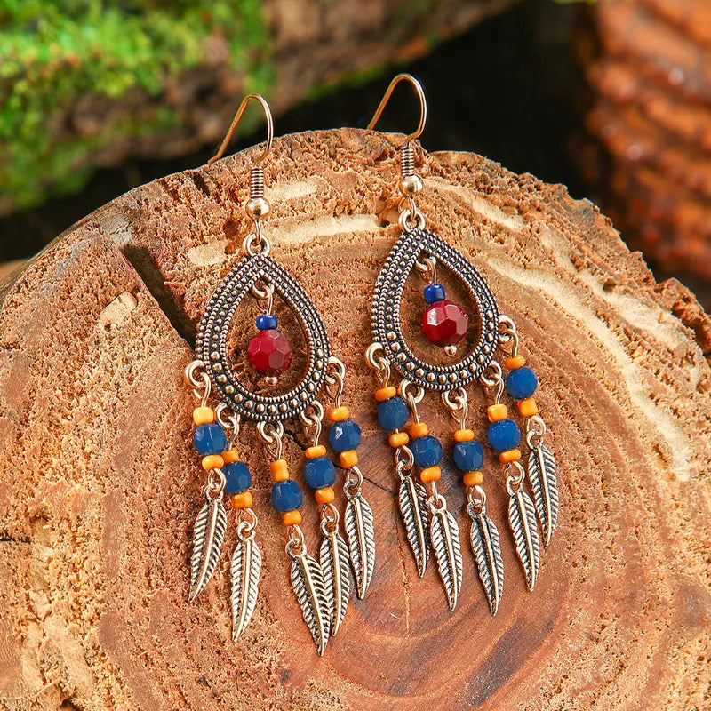 A pair of boho earrings with oxidised teardrop frames, blue and orange beads, and feather dangles, resting on a rustic wooden surface.