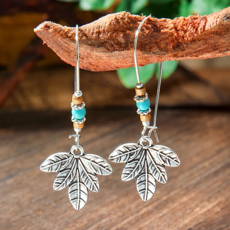 Close-up of boho earrings featuring silver leaf charms, turquoise beads, and wooden beads, hanging from a wooden branch.