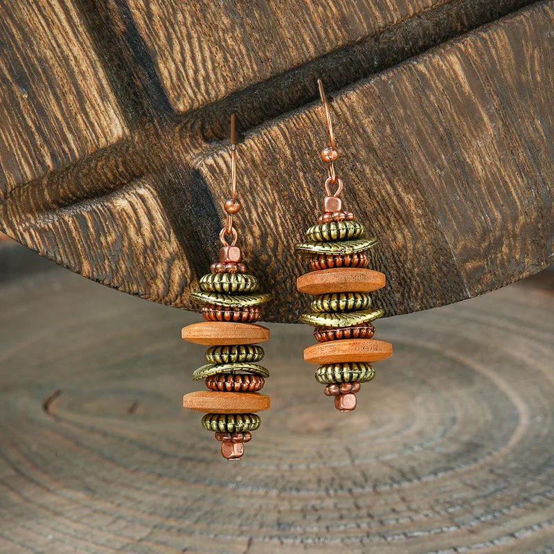 A pair of boho earrings with stacked wooden and metallic beads in brown, gold, and copper, hanging against a rustic wooden background.