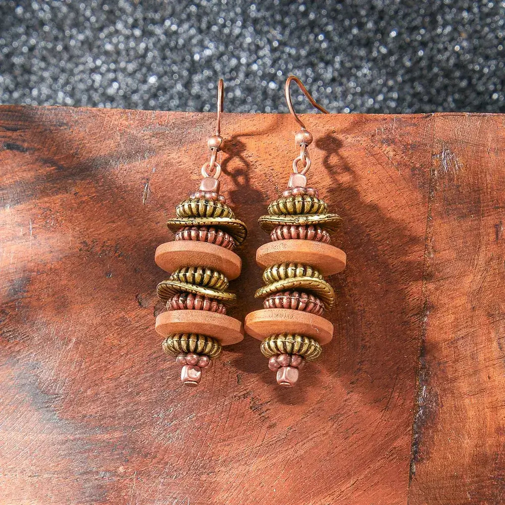 Close-up of a pair of boho earrings featuring stacked wooden and metallic beads in brown, gold, and copper, displayed on a wooden surface.