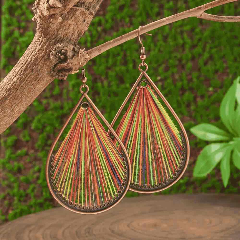 Close-up of a pair of multicolor threaded boho earrings hanging from a tree branch, with people blurred in the background.